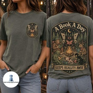 May include: An olive green t-shirt featuring a small circular graphic on the front chest and a large detailed illustration on the back. The back design depicts a moth, a sasquatch, a deer, and other creatures reading books by a campfire with the text A BOOK A DAY KEEPS REALITY AWAY.