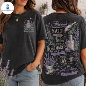 May include: A charcoal gray t-shirt featuring a small graphic on the front chest of a bottle and the text "Fall in love whenever you can." The back displays a large graphic with lavender, a bottle, and text: "Always throw spilled salt over your left shoulder. Keep rosemary by your garden gate. Plant lavender for luck. And fall in love whenever you can."