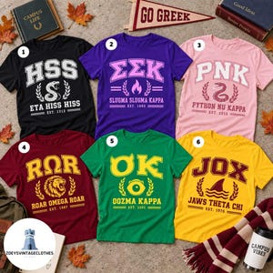 May include: Six short-sleeve crew neck t-shirts in black, purple, pink, maroon, green, and yellow. Each shirt features a unique Greek-style letter design, a central graphic icon, and text including names like Eta Hiss Hiss, Slugma Slugma Kappa, and Jaws Theta Chi.