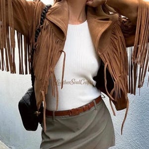 Op de afbeelding: Een bruin su&egrave;de jasje met lange franjes op de mouwen en voorpanden, gedragen over een wit geribbelde top en een grijze rok met een bruine leren riem. De tekst LeatherSoulCrafts is zichtbaar op de witte top.
