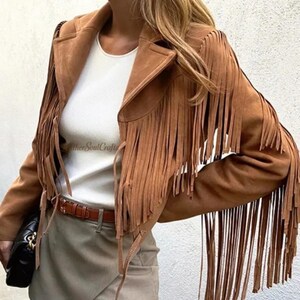 Op de afbeelding: Een bruin su&egrave;de jasje met lange franjes langs de mouwen en voorpanden, gedragen over een wit topje en een lichtgrijze rok met een bruine riem. Een zwarte handtas wordt aan de zijkant vastgehouden en de tekst LeatherSoulCrafts is zichtbaar.