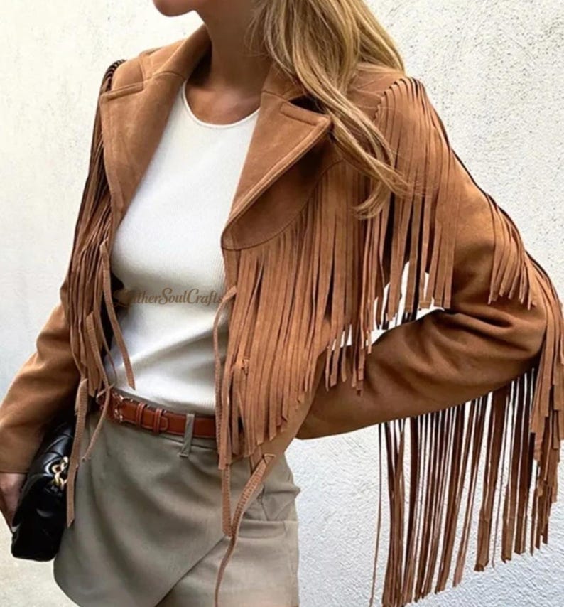Op de afbeelding: Een bruin su&egrave;de jasje met lange franjes langs de mouwen en voorpanden, gedragen over een wit topje en een lichtgrijze rok met een bruine riem. Een zwarte handtas wordt aan de zijkant vastgehouden en de tekst LeatherSoulCrafts is zichtbaar.