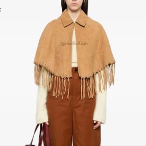 May include: A tan suede cape featuring a pointed collar, a front zipper closure, and long fringe detailing along the bottom hem. The garment is layered over a cream-colored long-sleeve top and brown trousers. The text LeatherSoulCrafts is visible.