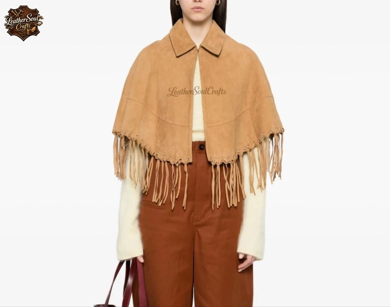 May include: A tan suede cape featuring a pointed collar, a front zipper closure, and long fringe detailing along the bottom hem. The garment is layered over a cream-colored long-sleeve top and brown trousers. The text LeatherSoulCrafts is visible.