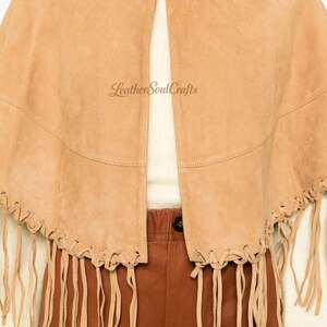 May include: A tan suede cape featuring a classic collar, a front opening, and long fringe detailing along the bottom hem. The text LeatherSoulCrafts is printed on the front. The garment is worn over a cream top and brown trousers.