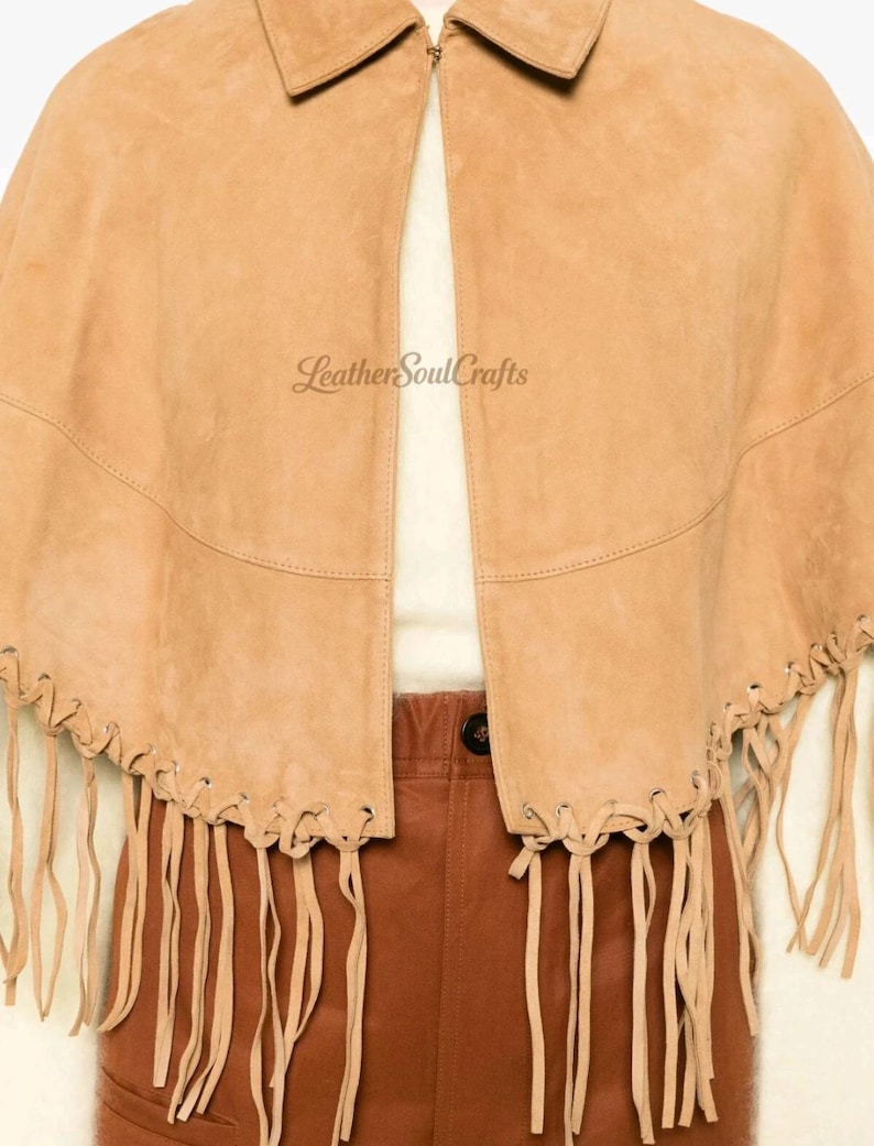 May include: A tan suede cape featuring a classic collar, a front opening, and long fringe detailing along the bottom hem. The text LeatherSoulCrafts is printed on the front. The garment is worn over a cream top and brown trousers.