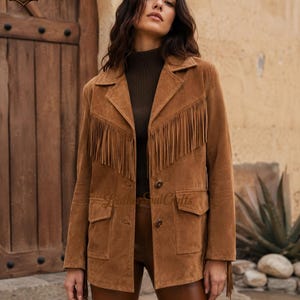 May include: A tan suede jacket featuring long fringe detailing across the chest, a notched lapel collar, two front flap pockets, and two brown buttons. The jacket has long sleeves with fringe accents at the cuffs and a structured, tailored fit.