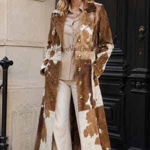 Puede incluir: Un abrigo largo de piel de vaca con un patrón de parches de pelo marrón y blanco. El abrigo tiene cuello de solapa, mangas largas y un cierre frontal con cinco botones dorados. El texto LeatherSoulCraft es visible en la imagen.