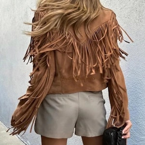Op de afbeelding: Een bruin su&egrave;de jasje met lange franjes op de rug, schouders en mouwen. Het jasje heeft een V-vormig rugpand en wordt gedragen met een lichtgrijze korte broek en een kleine zwarte handtas. De tekst LeatherSoulCrafts is zichtbaar.