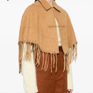 May include: A tan suede cape featuring a collared neckline and long fringe detailing along the bottom hem. The garment is accented with visible lacing along the lower edge and includes the text LeatherSoulCrafts printed on the fabric.