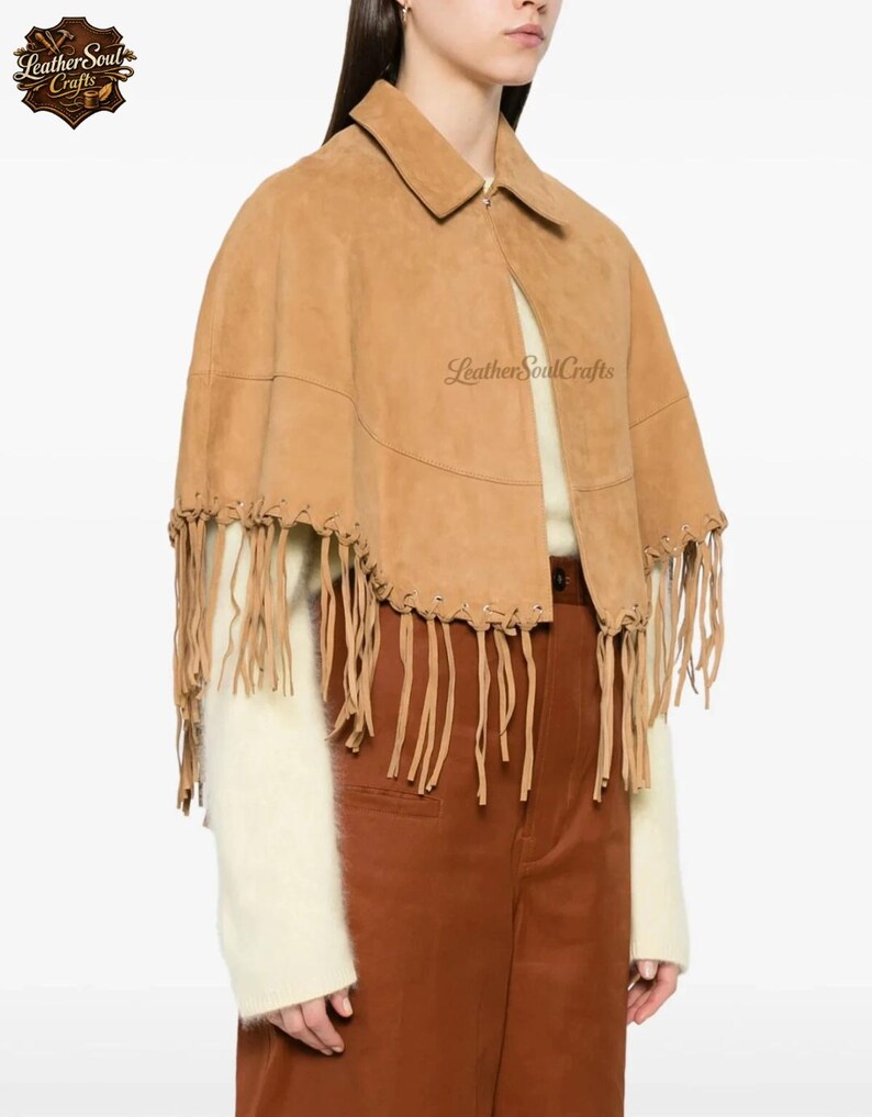 May include: A tan suede cape featuring a collared neckline and long fringe detailing along the bottom hem. The garment is accented with visible lacing along the lower edge and includes the text LeatherSoulCrafts printed on the fabric.