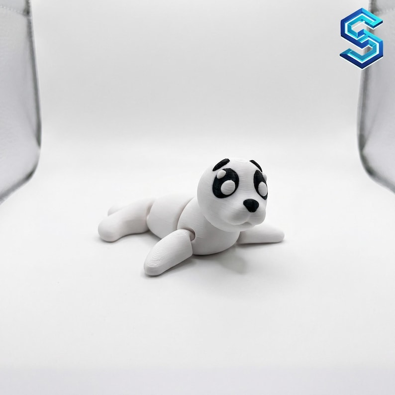 May include: A white 3D printed seal figurine with black painted eyes and a black nose. The seal is in a lying position with articulated joints visible at the neck and flippers, featuring a smooth matte finish and a rounded head shape.