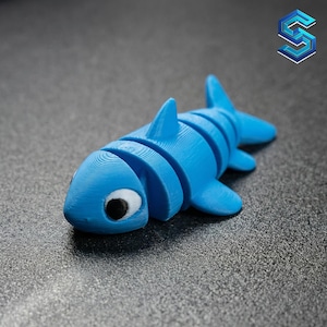 May include: A bright blue articulated shark figurine featuring a segmented body design, a prominent dorsal fin, and two side fins. The shark has a white eye patch with a black pupil, and the surface shows visible 3D printed layer lines throughout.