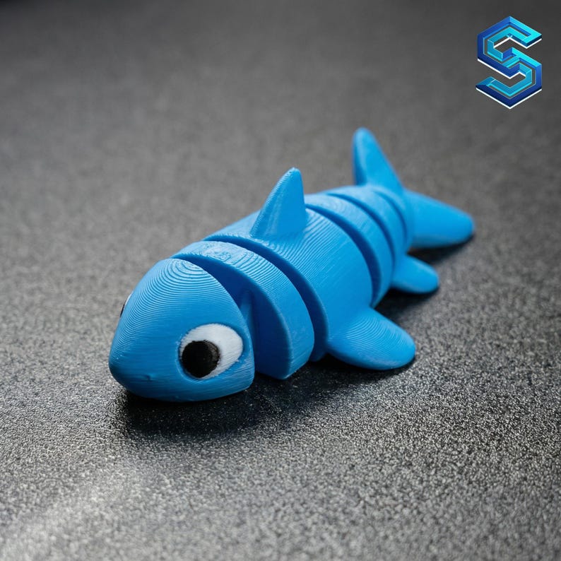 May include: A bright blue articulated shark figurine featuring a segmented body design, a prominent dorsal fin, and two side fins. The shark has a white eye patch with a black pupil, and the surface shows visible 3D printed layer lines throughout.