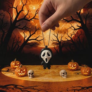 May include: A small black and white ghost-faced character keychain featuring a white mask with hollow eyes and an open mouth, attached to a metallic ball chain. The figure is suspended against a backdrop of a large orange moon and dark trees.