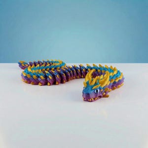 May include: A multi-colored articulated dragon figurine featuring a gradient of purple, blue, and teal scales. The dragon has prominent gold-colored horns and spikes along its back, with a long, segmented body that curves across the surface.