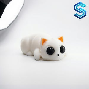 May include: A small, white, squishy cat figurine with a smooth, matte texture. The cat is in a lying-down position with large, round, solid black eyes, a tiny black nose, and bright orange inner ears, featuring a minimalist and rounded design.