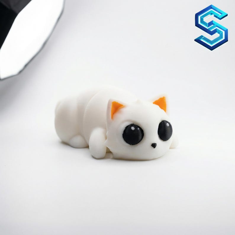 May include: A small, white, squishy cat figurine with a smooth, matte texture. The cat is in a lying-down position with large, round, solid black eyes, a tiny black nose, and bright orange inner ears, featuring a minimalist and rounded design.