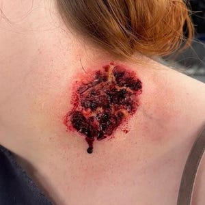 May include: A close-up of a realistic wound prosthetic applied to the skin. The wound is a deep red color with dark, clotted areas and a small, dark droplet. The skin around the wound is pale.