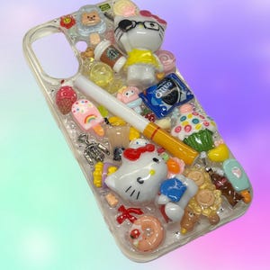 Pu&ograve; includere: Custodia trasparente per telefono decorata con vari ciondoli colorati, tra cui un personaggio di Hello Kitty, una sigaretta, un biscotto Oreo, un cupcake e una tazza di caff&egrave;. La custodia ha una variet&agrave; di altri piccoli oggetti decorativi.