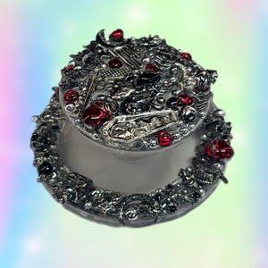 May include: Ornate silver-toned trinket box with a lid. The lid and base are heavily embellished with intricate designs, black accents, and red jewel-like embellishments. The box is set against a pastel gradient background.
