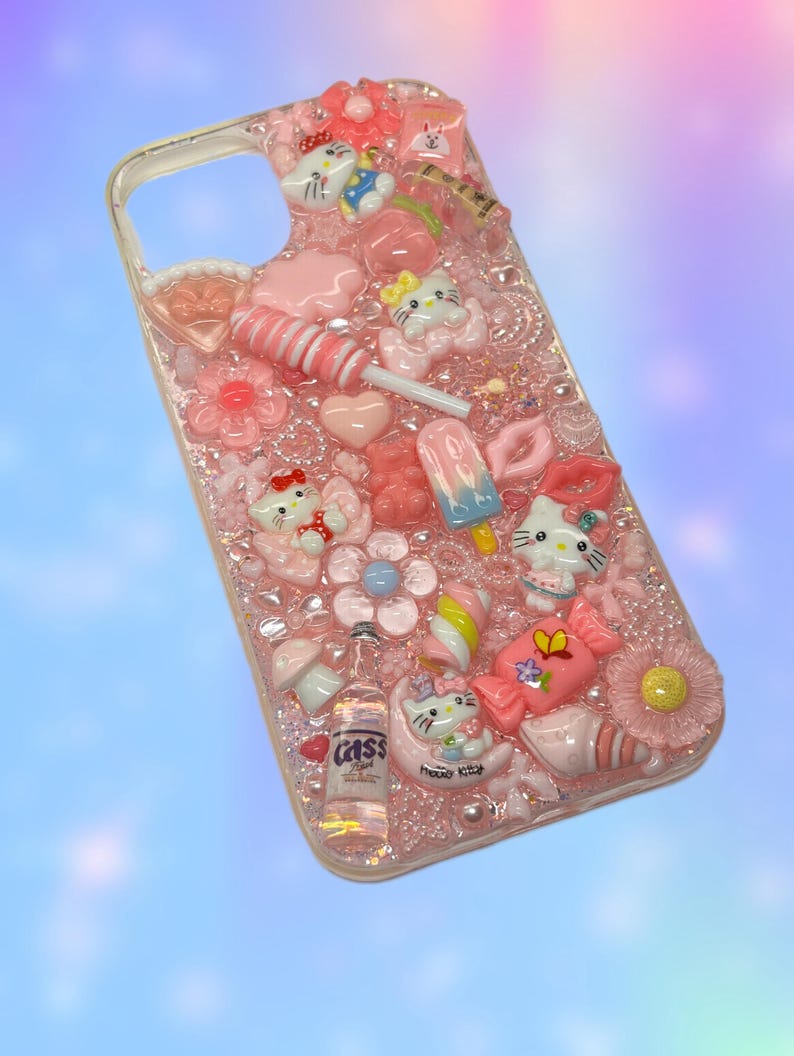 Może przedstawiać: R&oacute;żowe etui na telefon ozdobione r&oacute;żnymi zawieszkami, w tym kwiatami, sercami i figurkami Hello Kitty. Etui posiada r&oacute;wnież lizaka, małą butelkę piwa Cass i inne dekoracje o tematyce słodyczy.
