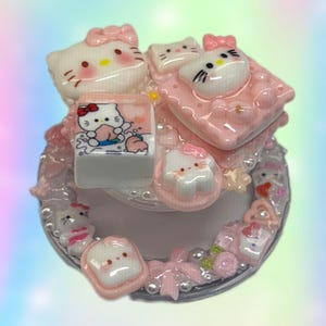 May include: A decorative, multi-layered object featuring Hello Kitty-themed embellishments. The design includes white and pink elements, such as Hello Kitty faces, bows, and small square blocks with illustrations. The base is clear with pearl-like accents.