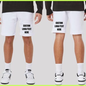 Puede incluir: Pantalones cortos deportivos blancos con el texto "CUSTOM LOGO/TEXT HERE" impreso en la parte delantera. Los pantalones cortos se muestran en una persona que usa zapatillas y calcetines blancos. La persona lleva una camisa negra de manga larga.