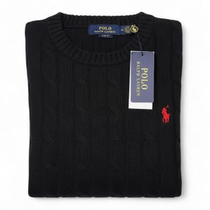 Puede incluir: Jersey negro de punto trenzado con cuello redondo. El jersey presenta un logotipo de polo rojo en el pecho y una etiqueta con el nombre de la marca. El jersey está doblado cuidadosamente sobre una superficie blanca.