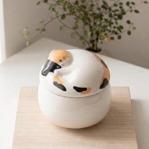 May include: A white ceramic jar with a lid shaped like a curled-up calico cat. The cat design features orange, black, and white markings. The jar sits on a light brown wooden surface, with a green plant in the background.