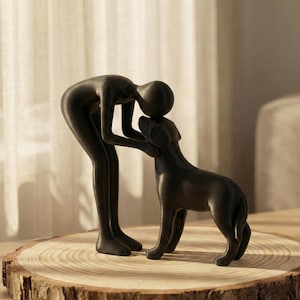 Peut inclure: Sculpture noire représentant une personne embrassant un chien. Le chien est debout, face à la personne. La sculpture est posée sur une tranche de bois, avec un grain naturel.