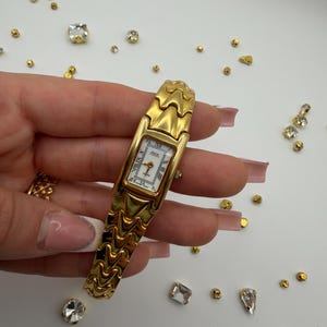 May include: Gold-toned wristwatch with a rectangular white face and Roman numeral hour markers. The watch has a textured, wavy band. Small gold and clear gemstones are scattered around the watch.