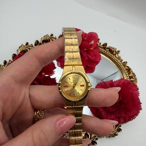 May include: A gold-toned wristwatch with a rectangular link band and a round face. The watch face is gold with red accents. The watch is held in front of a gold-framed mirror with red flowers.