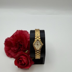 Op de afbeelding: Een goudkleurig polshorloge met een ruitvormige wijzerplaat en een getextureerde band. Het horloge wordt getoond op een zwarte standaard, met rode bloemen op de achtergrond. De wijzerplaat heeft de merknaam "Titan".