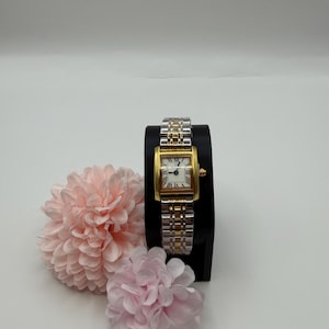 May include: A rectangular gold and silver wristwatch with a white face and Roman numerals. The watch is displayed on a black stand, next to two pink fabric flowers. The watch band is a combination of gold and silver links.