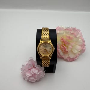 Op de afbeelding: Een goudkleurig Lorus-polshorloge met een ronde wijzerplaat, gouden wijzers en een datumvenster op de drie-uurpositie. Het horloge heeft een gestructureerde goudkleurige metalen schakelarmband en staat op een zwarte standaard naast twee roze bloemen.