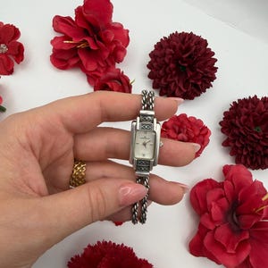 Puede incluir: Un reloj de pulsera rectangular plateado con esfera blanca y correa de cadena plateada. El reloj es sostenido por una mano, rodeado de flores rojas sobre un fondo blanco.