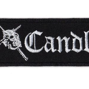 Może przedstawiać: Prostokątna czarna haftowana naszywka z białym napisem Candlemass gotycką czcionką. Po lewej stronie tekstu znajduje się biały haftowany wzór czaszki umieszczonej na dwóch skrzyżowanych mieczach, całość na czarnym tle z tkaniny.