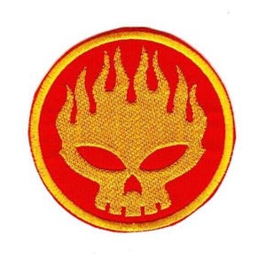 Puede incluir: Un parche bordado circular que presenta una calavera amarilla estilizada con cabello en forma de llamas sobre un fondo rojo sólido. El diseño está encerrado dentro de un borde amarillo grueso, con todo el emblema cosido en hilo vibrante.