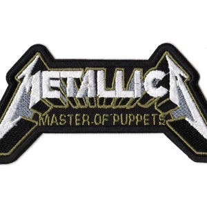 May include: An embroidered fabric patch featuring the Metallica logo in white with black and grey shading. Below the logo, the text MASTER OF PUPPETS is stitched in a gold-colored thread. The entire design is outlined with a thick black border.