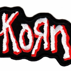 May include: An embroidered patch featuring the word Korn in a stylized white font with a red outline, set against a solid black background. The lettering has a jagged, hand-drawn appearance with thick, textured stitching throughout the design.