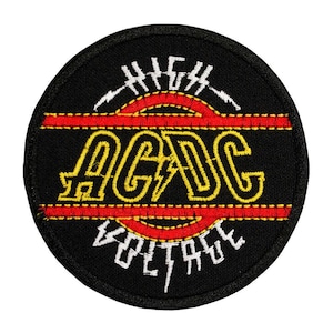 May include: A circular embroidered patch with a black background. It features the yellow text AC/DC in the center, flanked by two horizontal red lines. Above and below the text, the words HIGH and VOLTAGE are stitched in white thread.
