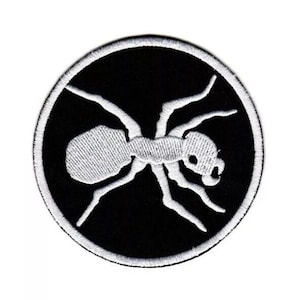 May include: A circular embroidered patch featuring a white ant silhouette centered on a solid black background. The design is enclosed by a thick white stitched border, creating a high-contrast graphic element suitable for fabric application.