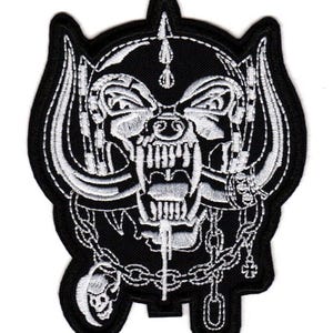 Puede incluir: Un parche bordado en blanco y negro que presenta una criatura estilizada con grandes cuernos curvados, dientes descubiertos y una cadena drapeada alrededor de la barbilla. El diseño incluye un pequeño dije de calavera y una cruz colgando.