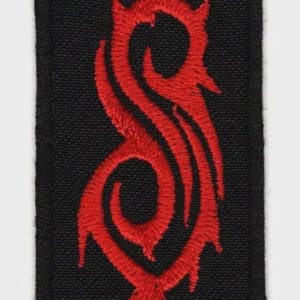 May include: A rectangular black fabric patch featuring a centred, vertically oriented, red embroidered tribal-style S-shaped logo. The design has sharp, pointed edges and a textured stitch pattern, with a solid black border framing the entire piece.