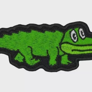 Puede incluir: Parche bordado con un cocodrilo verde de dibujos animados con ojos blancos y un contorno negro. El cocodrilo está caminando y tiene una expresión amigable. Un accesorio divertido para ropa o bolsos.