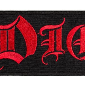 Könnte beinhalten: Ein rechteckiger bestickter Aufnäher mit dem Wort DIO in fetter, roter Schrift im gotischen Stil. Der Text ist auf einen einfarbigen schwarzen Stoffhintergrund gestickt, wobei eine passende schwarze Umrandung die gesamte rechteckige Form einfasst.