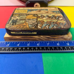 May include: A rectangular wooden block featuring a comic book panel titled ATTACK OF THE FLYING GORILLAS and COMIC MAGAZINE. The block measures approximately 6 inches in width as shown by the blue ruler placed directly in front of the object.