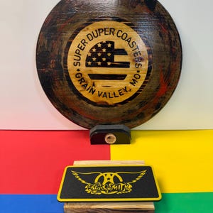 May include: A round wooden coaster with a dark stained finish and a central graphic featuring an American flag and the text 'SUPER DUPER COASTERS GRAIN VALLEY, MO'. Below it, a rectangular black coaster displays a yellow Aerosmith logo.