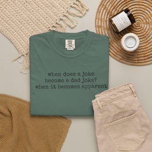 May include: A sage green short-sleeved t-shirt with a crew neckline and a Comfort Colors brand label at the collar. The shirt features black typewriter-style text that reads: when does a joke become a dad joke? when it becomes apparent.